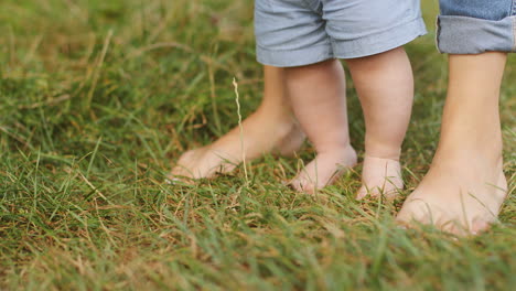 close up of a baby boy taking his first steps in the grass while holding his mother's hands 1