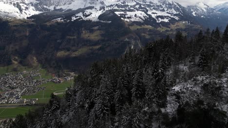 An-aerial-view-captures-snow-covered-forests-atop-the-hills-above-Glarus,-a-small-town-nestled-in-the-Swiss-Alps,-blending-natural-splendor-with-urban-residential-charm