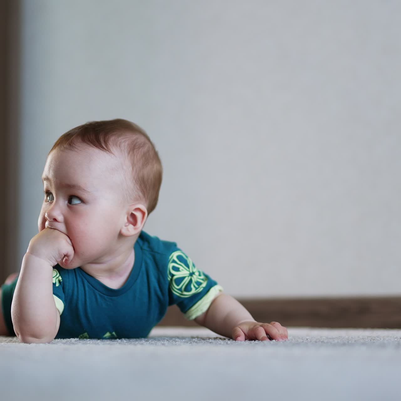 Little Caucasian infant boy lies on the floor banging hands and feet upon it. Cute emotional toddler takes a fist into mouth looking back