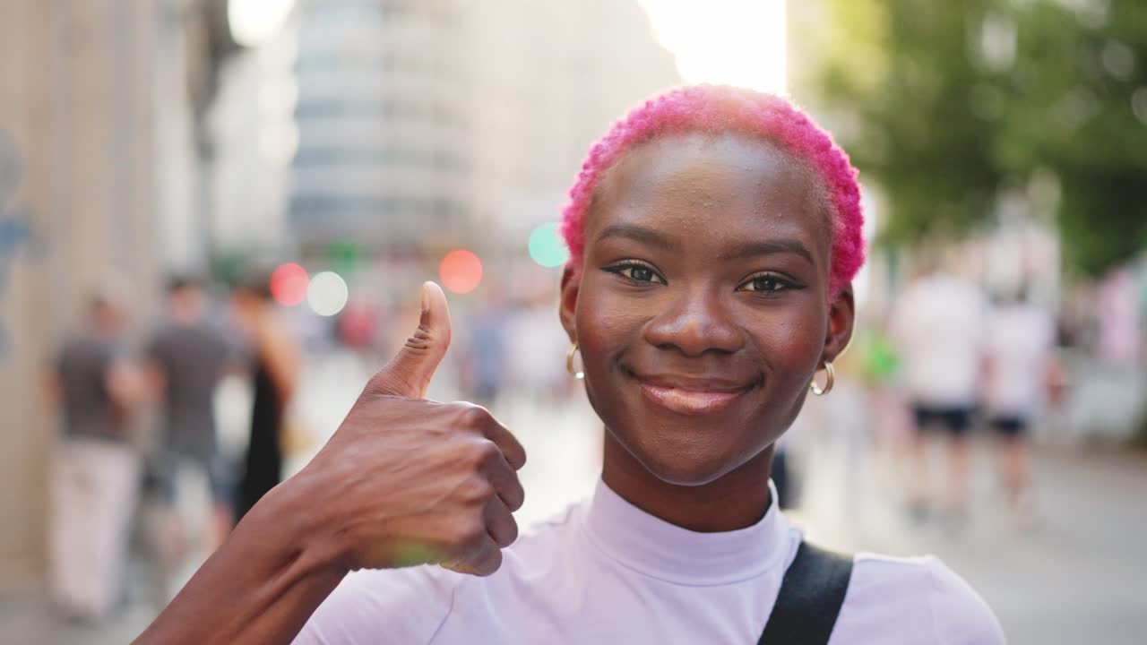 Una giovane donna di colore con i capelli rosa che sorride e fa il pollice in su in un contesto urbano