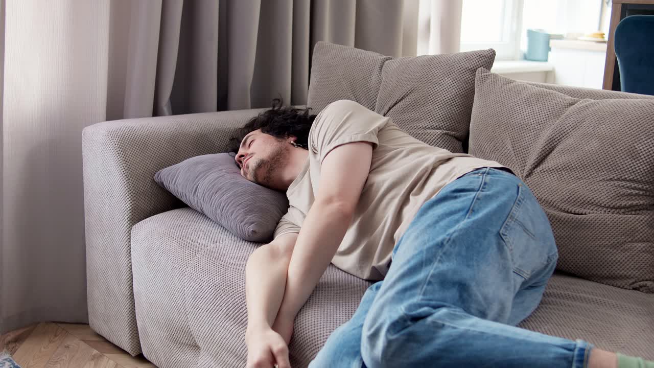 가운 갈색 셔츠를 입고 곱슬머리를 가진 피곤한 갈색 남자가 현대 아파트의 소파에 누워있는 동안 베개에 떨어집니다.