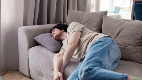 Ein-Müder-Brünetter-Typ-Mit-Lockigem-Haar-In-Einem-Hellbraunen-Hemd-Fällt-Auf-Ein-Kissen,-Während-Er-Auf-Dem-Sofa-In-Einem-Modernen-Apartment-Liegt