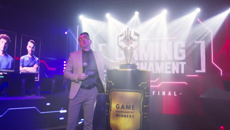 male announcer showing gaming tournament trophy man host with microphone walking and talking to audience while demonstrating golden cup on stand during esports championship