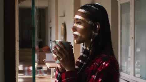 Mixed-race-woman-standing-by-window-at-home-having-coffee