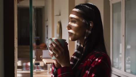 mujer de raza mixta de pie junto a la ventana en casa tomando café