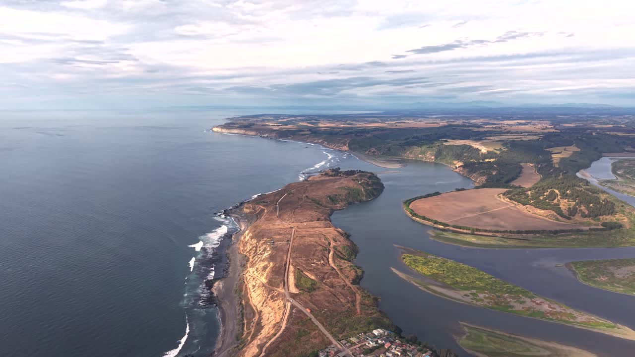 Toma cinematográfica aérea con dron del estuario de un río ancho que desemboca en el Océano Pacífico, mostrando un paisaje costero espectacular con acantilados secos, una península de arena y olas rompiendo en Chile y en 4K