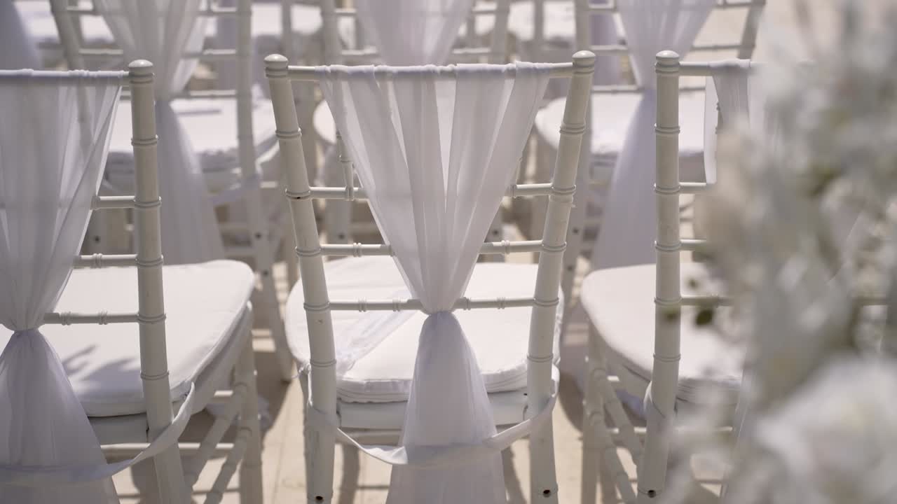 Filas de sillas blancas están dispuestas para una ceremonia de boda en un lugar al aire libre. Unas telas suaves añaden un toque especial para los invitados que asisten a esta alegre celebración.