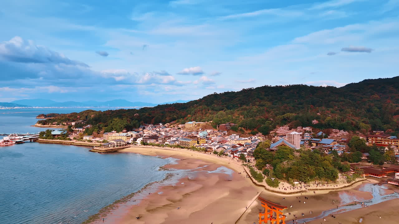 Low tide at the Seto Inland Sea allowing tourists walk around the famous landmark of red gates. Drone footage approaching inhabited area and mountains near Itsukushima Shrine, Japan.
