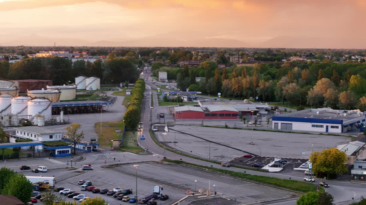 Aerial panning drone shot reveals the Cremona refinery with large fuel storage tanks, industrial infrastructure, roads, and parking lots under warm golden hour light in Lombardy, Italy
