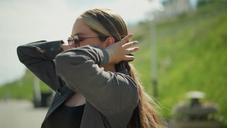 lady wearing a black top under a grey jacket and sunglasses, adjusting her hair with a focused expression, the setting is outdoors, with blurred greenery in the background on a bright day