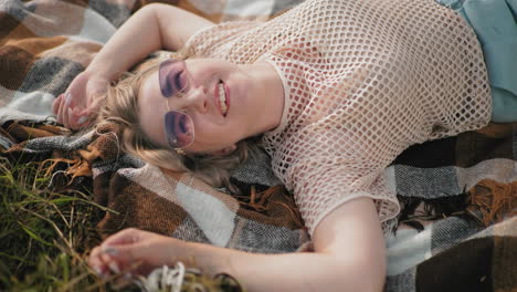 woman in net top and sunglasses lying on checkered blanket in grassy field, smiling joyfully while touching grass, relaxed outdoor moment with soft sunlight