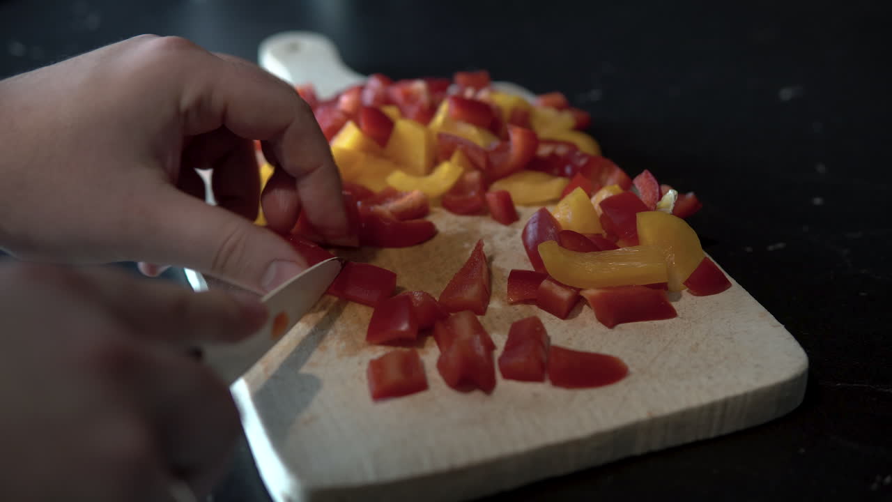 Sliced red peppers on wood board on black worktop, close-up for hands and knife