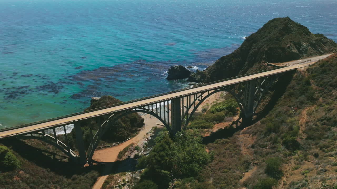 Sightly arched bridge for transport between the mountains on the coast of Pacific. Beautiful blue water sparkling in the sun at backdrop.