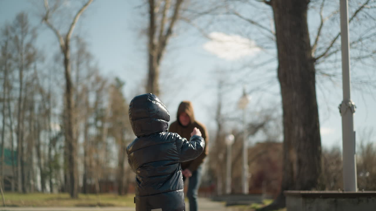 A close-up of a father and son playfully jogging together on a park pavement, both dressed in jackets, jeans, and boots, the father playfully holds his son amidst trees and lamp posts lining