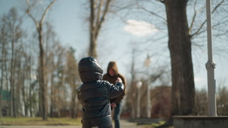 a close-up of a father and son playfully jogging together on a park pavement, both dressed in jackets, jeans, and boots, the father playfully holds his son amidst trees and lamp posts lining