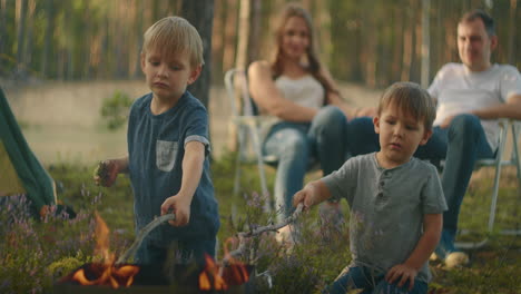 Two-boys-sitting-by-a-fire-against-a-tent-in-the-woods-on-the-shore-of-the-lake-fry-marshmallows-on-fire.-Brothers-3-6-years-together-burn-sticks-on-fire