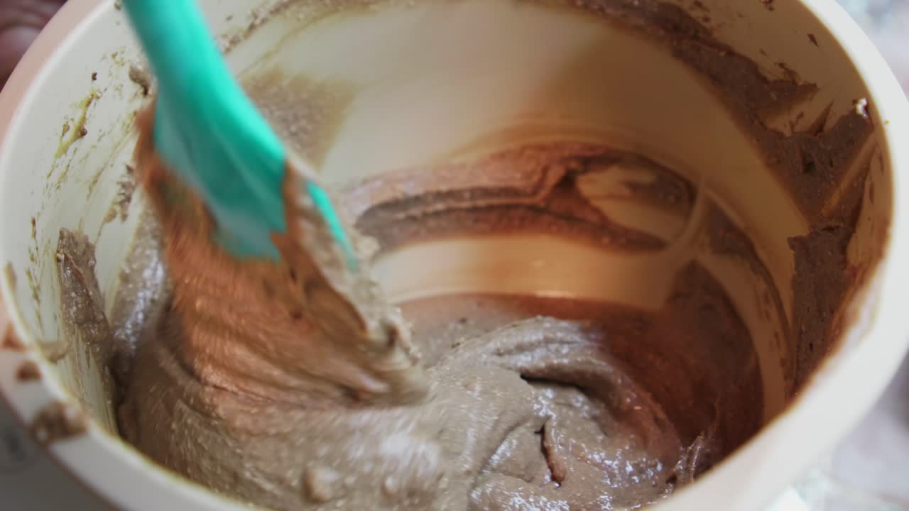 Hands mix cake batter in a bowl at home kitchen. A spatula stirs through the thick mixture. The process shows how ingredients combine. The kitchen is well-lit, showing the mixing action
