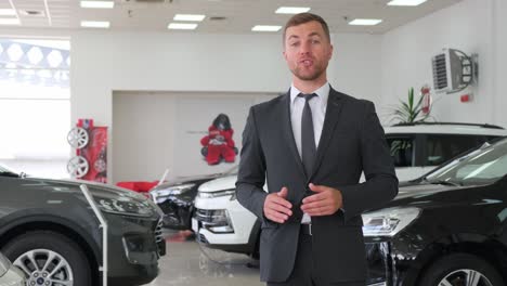 man in suit trousers gesturing in front of car showroom. a man in a suit and tie is gesturing towards the cars in front of the car showroom, showcasing the latest models with shiny tires and wheels for the ultimate automotive experience