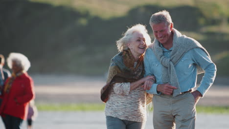 Amante-Pareja-Senior-Activa-Caminar-Del-Brazo-A-Lo-Largo-De-La-Costa-En-Vacaciones-De-Invierno-En-La-Playa