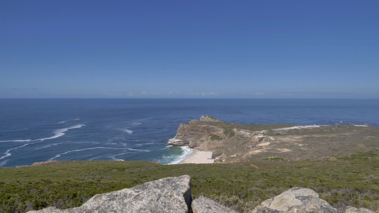 Vidstrakt udsigt over barske klipper og en sandstrand, der grænser op til det dybblå hav med blide bølger og hvide skumspor ved Cape Point, Cape Peninsula, Sydafrika