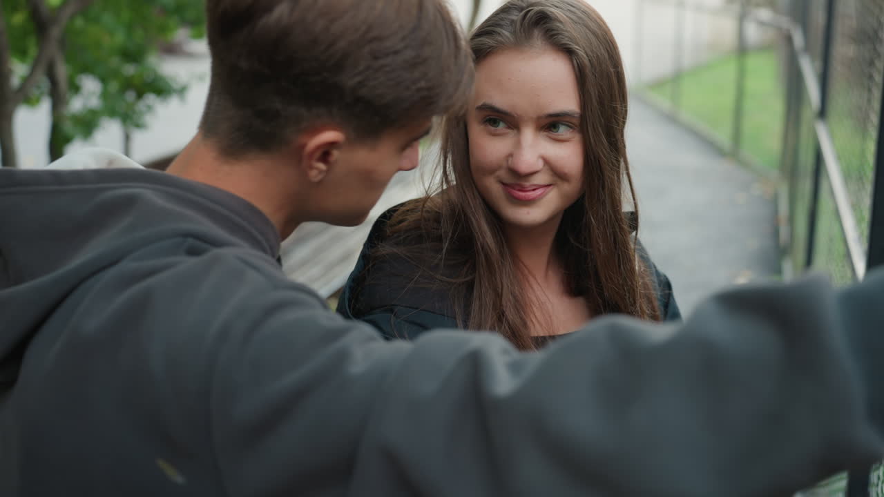Kaukasisk tenåringspar deler smil på telefonen, gutten lener seg nærmere for å se skjermen, jenta smiler forsiktig, intimt ærlig portrett langs parkstien, mykt naturlig lys og varm tone, leken flørting