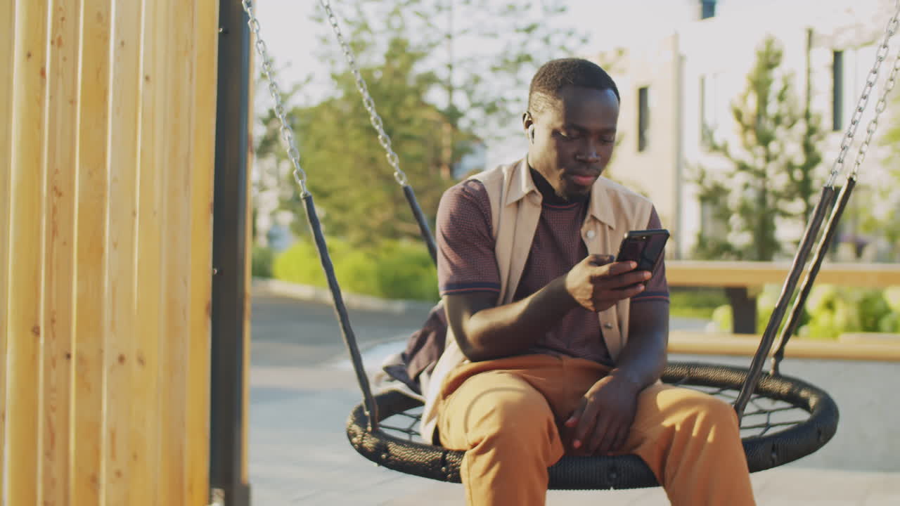 Black Man Sitting Using Smartphone on Swing Outdoors