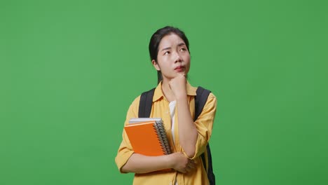 asian woman student with a backpack and some books thinking and looking around while standing in the green screen background studio