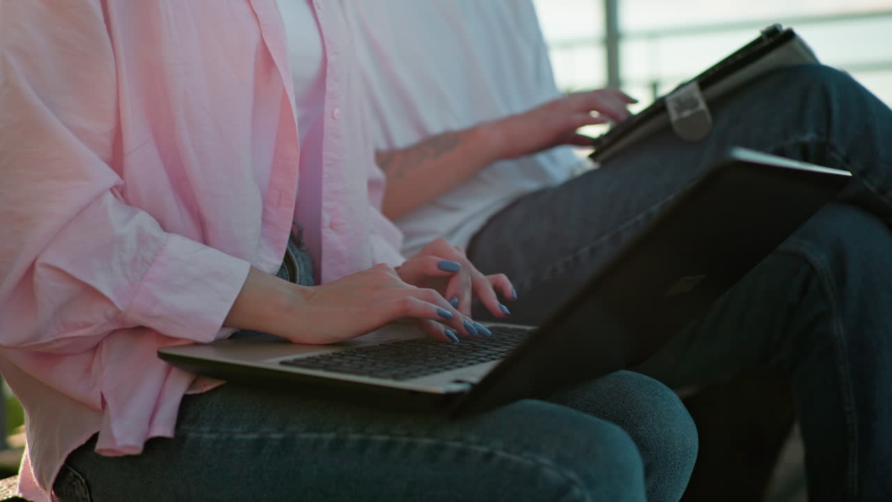 핑크 과 청바지를 입은 젊은 여성이 야외에 앉아있는 동안 파란 손을 가진 노트북에서 타이핑하는 클로즈업, 다른 사람 에는 또한 장치에서 타이핑에 집중합니다.