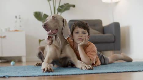 niño pensativo tirado en el suelo y acariciando a su gran perro lindo en casa y mirando la cámara