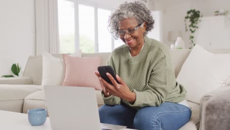 Portrait-of-happy-senior-african-american-woman-sitting-on-sofa,-using-laptop-and-smartphone