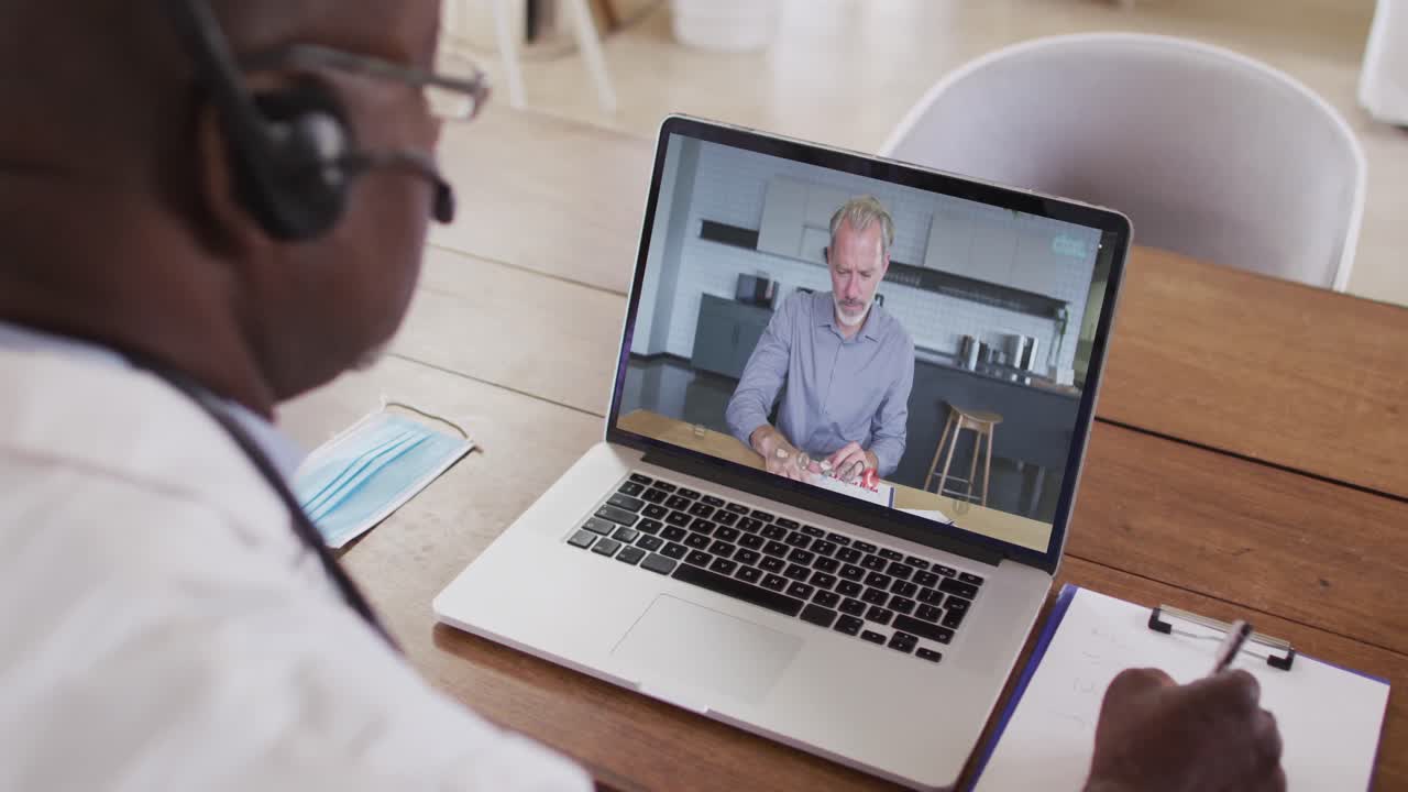 African american male doctor wearing phone headset taking notes while having a video call on laptop