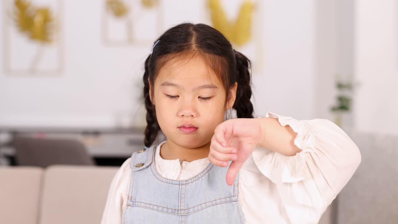 Young Asian girl in casual clothing gives a thumbs down gesture, then smiles, in a bright living room with soft natural lighting and static camera