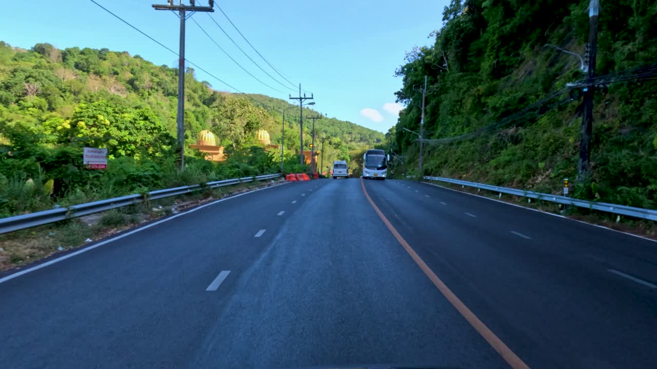 Vehicles travel on sunlit, winding mountain road with lush greenery and urban traffic in Phuket
