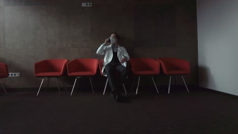 a young doctor in a white work coat and respirator sits in a dark room and consults over the phone
