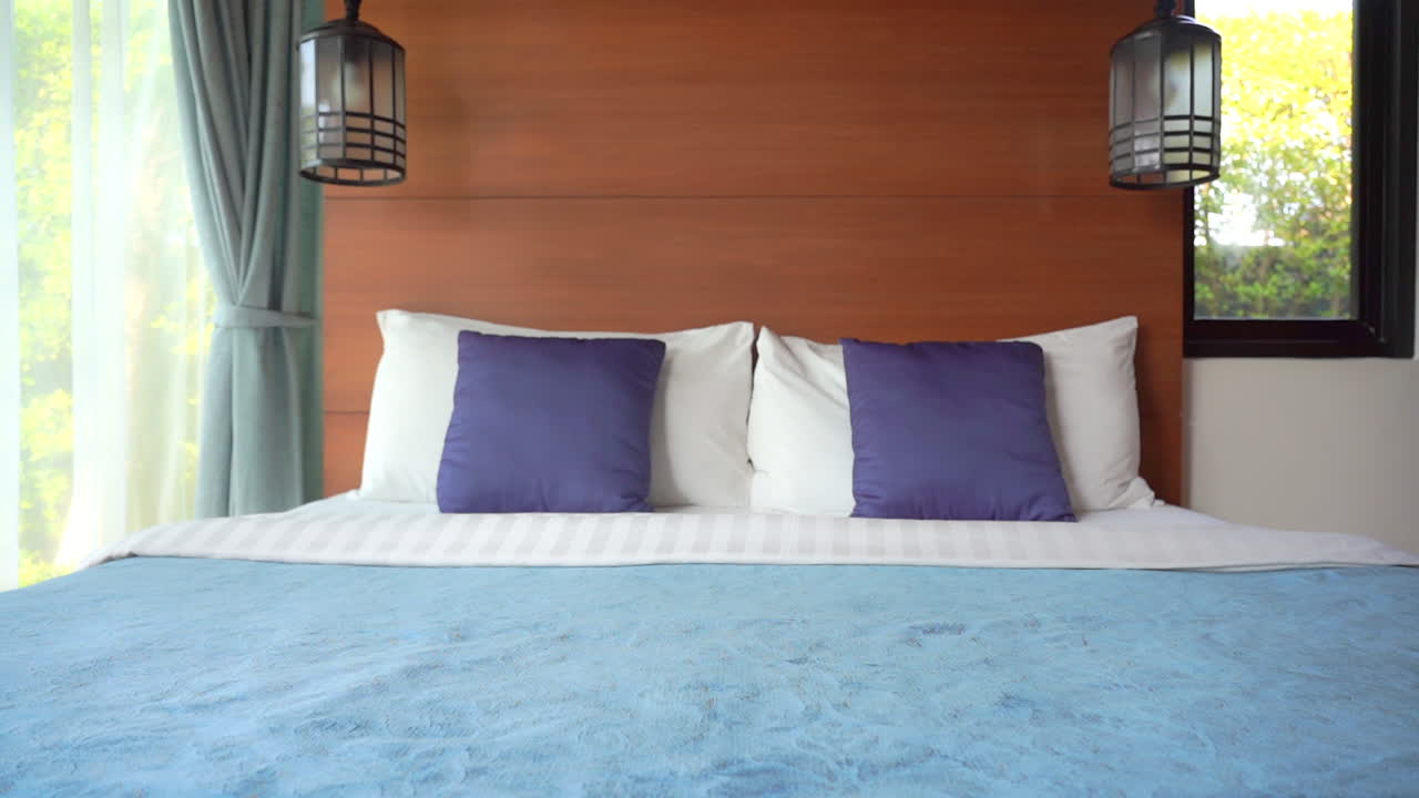 Panning shot hotel bedroom clean made up bed, blue pillows and blanket wooden wall, Morning lights