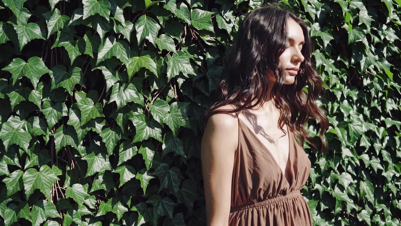 Young woman with long brown hair wearing a summer dress is posing near a green ivy wall, creating a harmonious blend of natural beauty and fashion