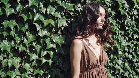 young woman with long brown hair wearing a summer dress is posing near a green ivy wall, creating a harmonious blend of natural beauty and fashion