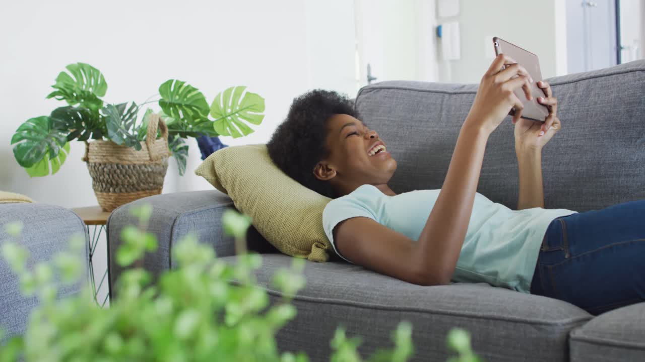 una adolescente afroamericana feliz acostada en el sofá, usando una tableta