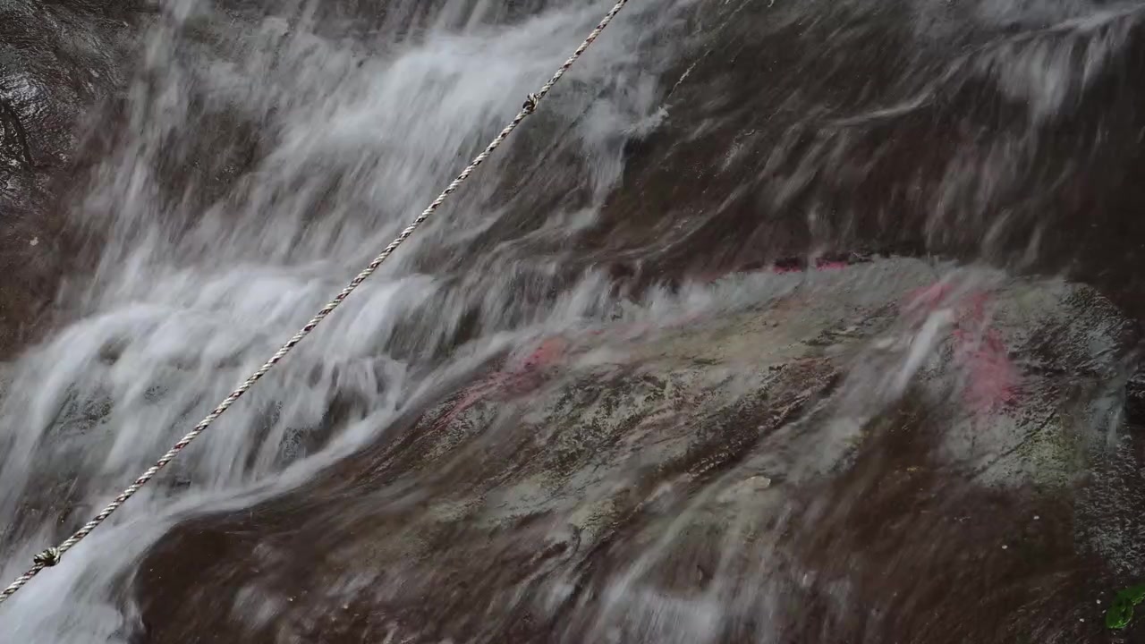 Una corda attraversa diagonalmente una cascata che scende su rocce scure e texturizzate con striature rosse e rosa