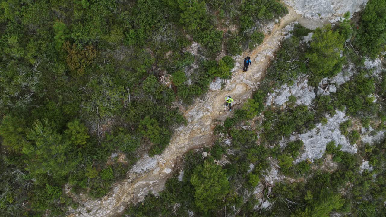 Two Hikers Trekking On Narrow Rocky Trail Through Dense Green Shrubs And Rugged Terrain In Noli, Liguria, Italy. aerial topdown shot