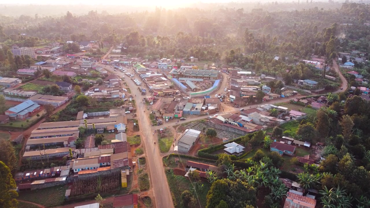 Filmisk luftfoto af en travl landlig markedsby i Embu, Kenya, der viser butikker, veje, hjem, køretøjer og omkringliggende landbrugsjord i varmt aftenlys