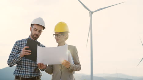 caucasian man and woman engineers wearing a helmet watching some blueprints and using tablet while talking at wind station of renewable energy