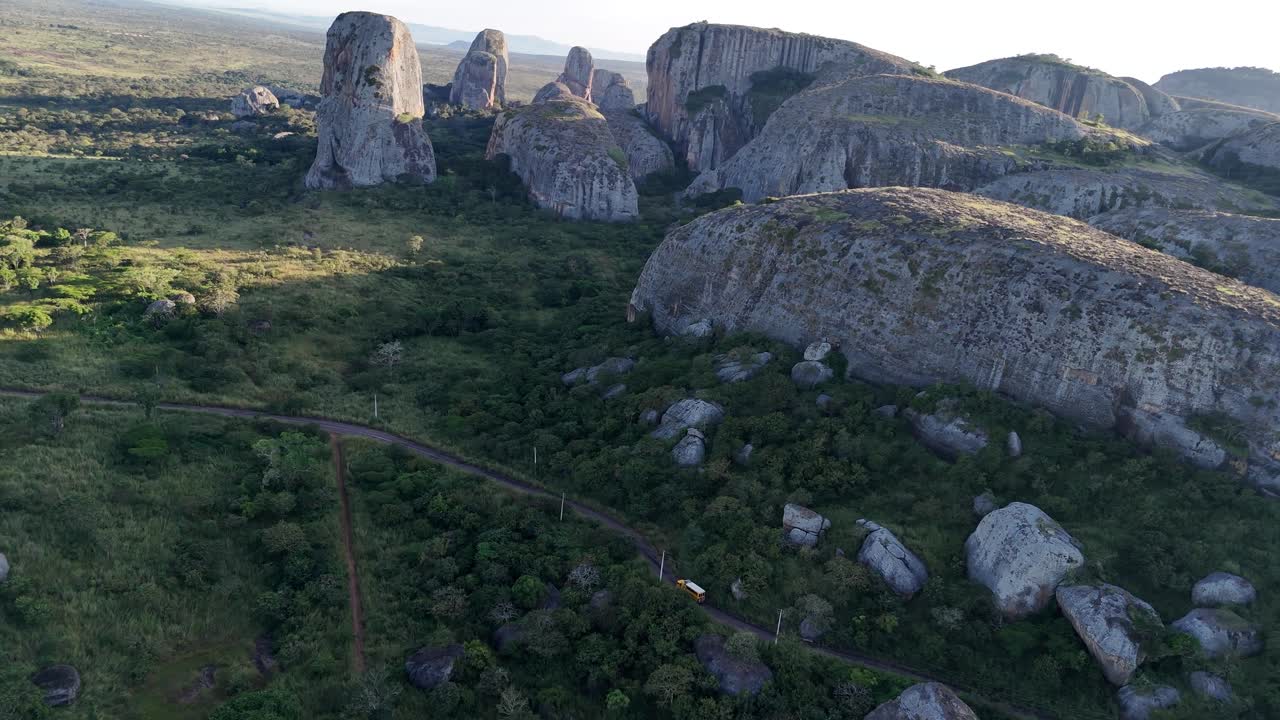 Vuelo cinematográfico en dron a baja altura de las antiguas rocas negras de Pungo Andongo, Angola; camioneta camper amarilla viajando por África en una carretera estrecha entre la formación rocosa, vista aérea panorámica