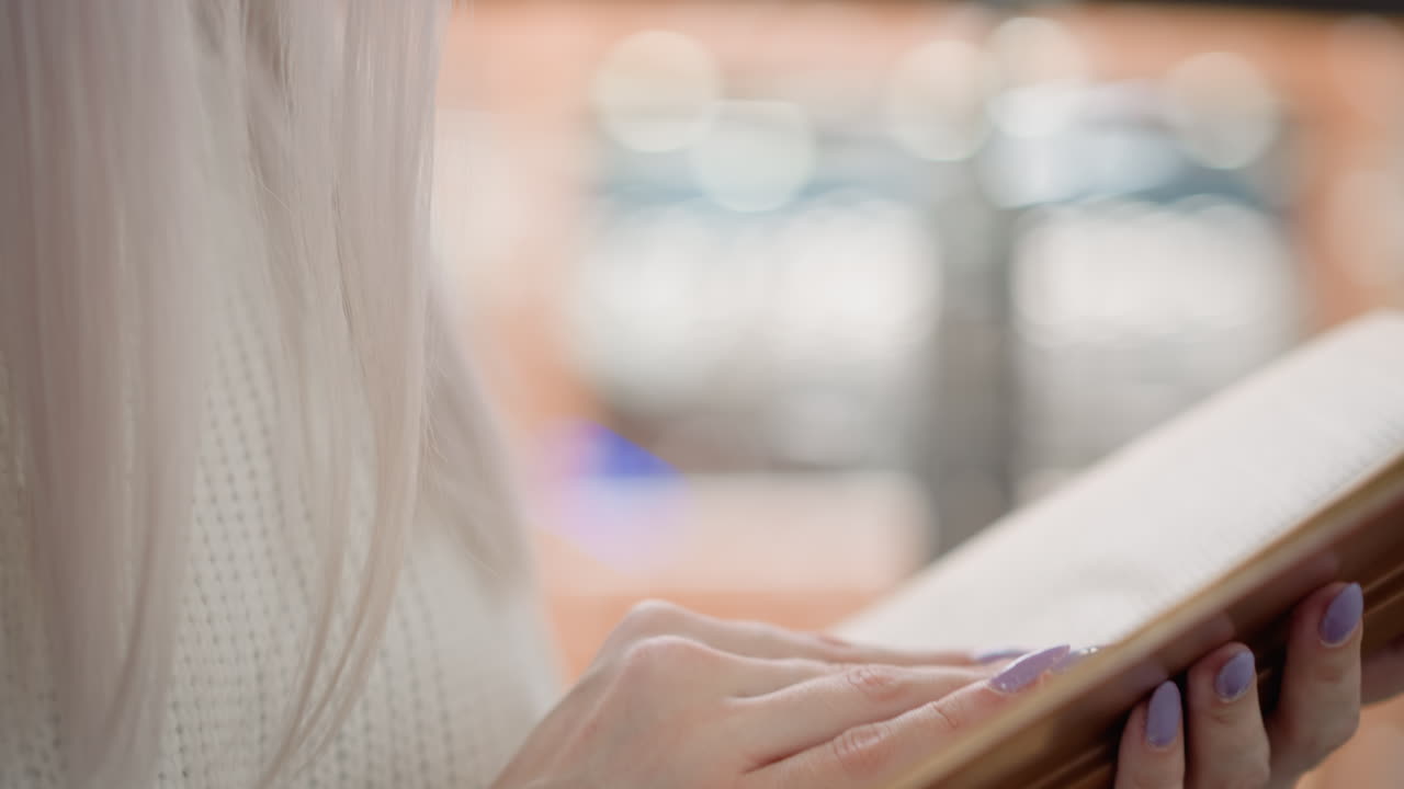 close up shot of mindful woman from head to hands holding book reading and gently flipping pages while seated on bench in shopping mall soft bokeh background warm indoor glow calm study moment