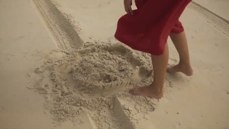 una mujer con un vestido rojo se para en la arena luego se inclina agarra la arena en sus manos y juega con ella