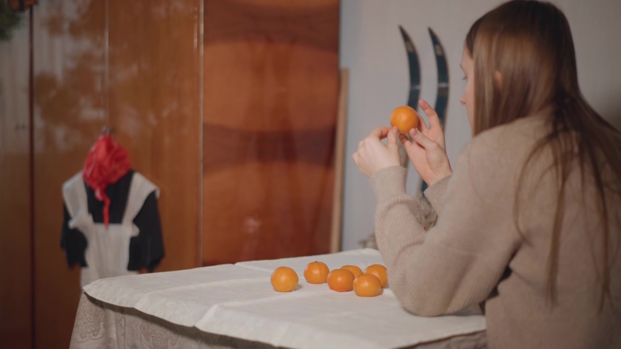 Pige skræller citrusfrugt. Kvinde i vintage tøj skræller mandarin. Ung kvinde skræller appelsin i retro omgivelser. Ung dame i vintage tøj skræller roligt en mandarin ved sit bord.