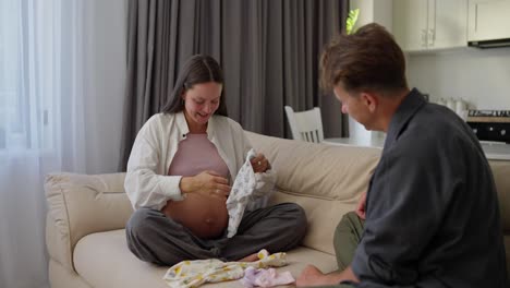 happy brunette pregnant woman tries on clothes for her baby whose child they are expecting together with her husband at home on the sofa