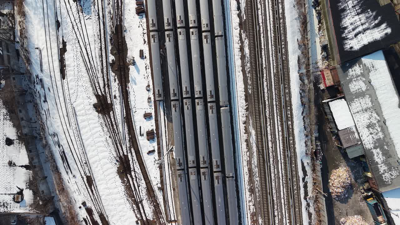 Top down aerial perspective flying over a large rail yard with multiple passenger trains parked on snowy tracks during winter, showcasing the complex network of railway lines and switches