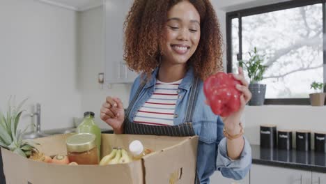Happy biracial woman unpacking groceries from box with recycle text in kitchen, slow motion