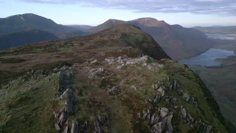 Hiker-on-distant-rocky-outcrop-walking-down-from-summit-with-orbit-revealing-deep-valley-with-lake-Buttermere